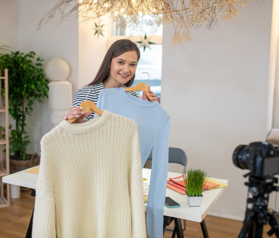 What to choose. Pleasant nice woman standing in front of the camera while holding two pieces of clothes