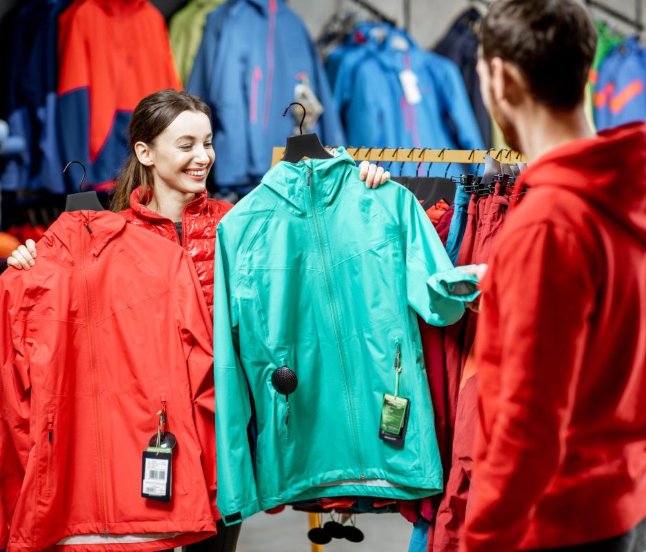 Young woman choosing between red and turquoise jacket standing with boyfriend in the sports shop